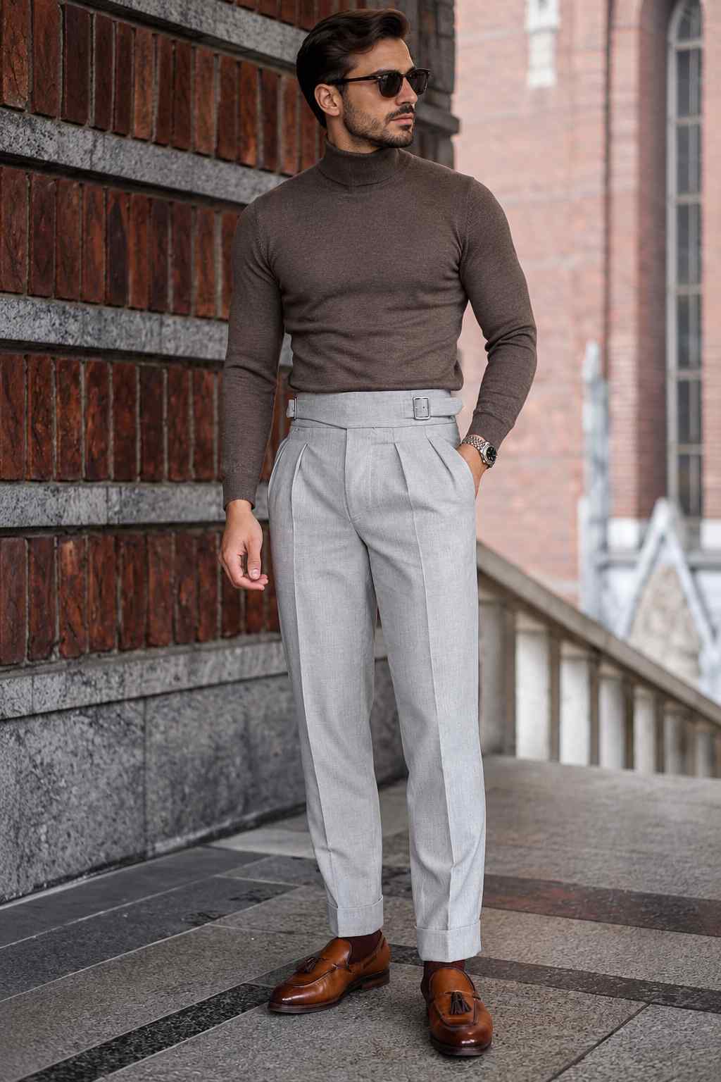 Men's grey high-waisted Gurkha trousers with double pleats, adjustable side buckle, tailored straight legs and turn-up cuffs, styled with brown loafers.