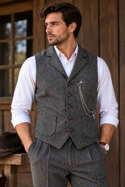 Man wearing a grey herringbone tweed waistcoat with matching trousers, a white shirt, brown buttons, and a decorative pocket chain against a rustic wooden background.
