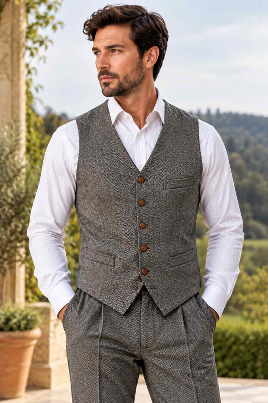 Man wearing a grey herringbone tweed waistcoat with brown buttons and front pockets over a white dress shirt, styled with matching patterned trousers.