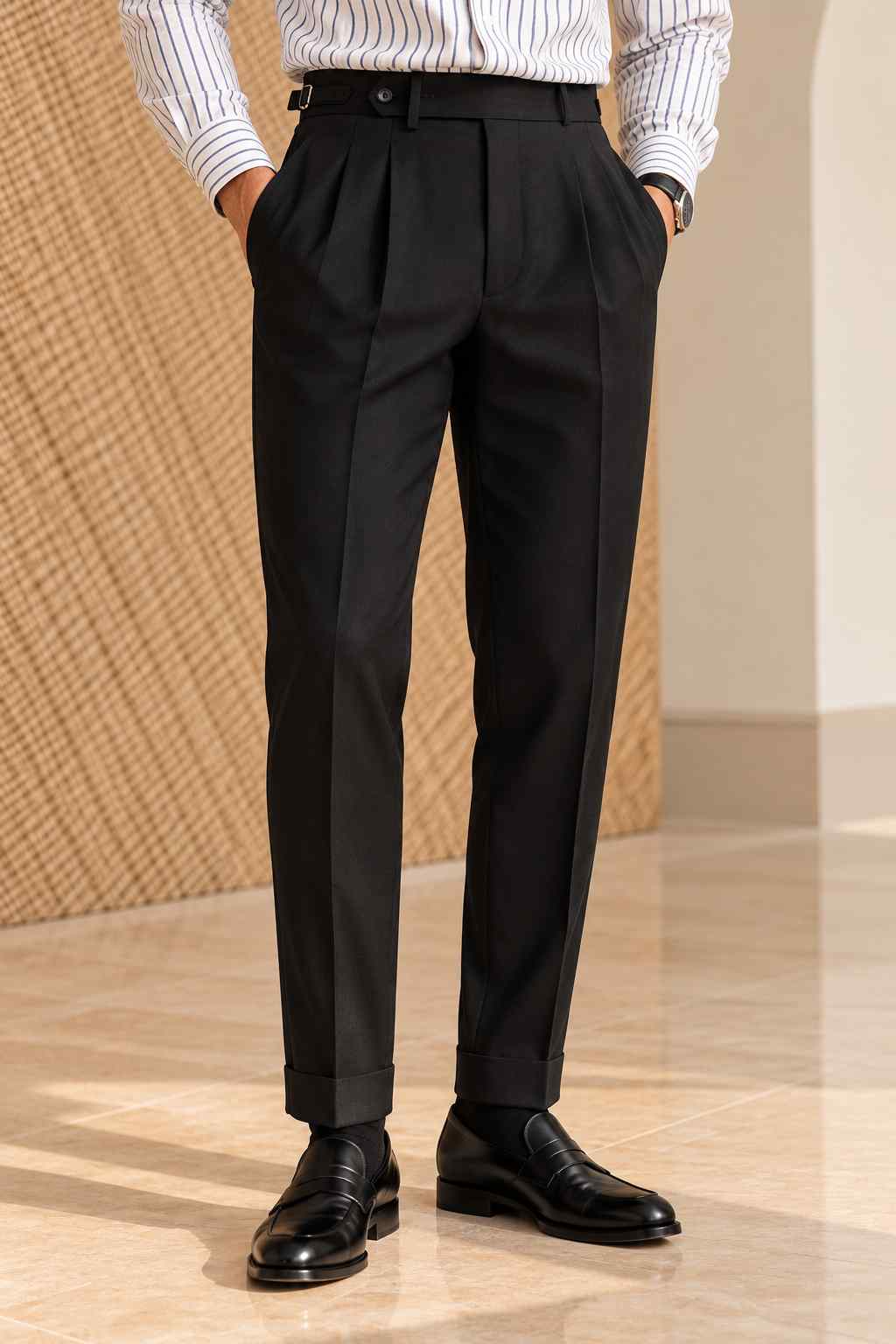 Men's black high-waisted pleated trousers with a tailored fit, belt fastening and cuffed hems, styled with a white striped shirt and black loafers.