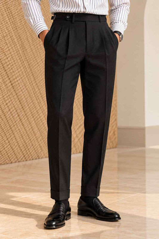 Men's black high-waisted pleated trousers with a tailored fit, belt fastening and cuffed hems, styled with a white striped shirt and black loafers.
