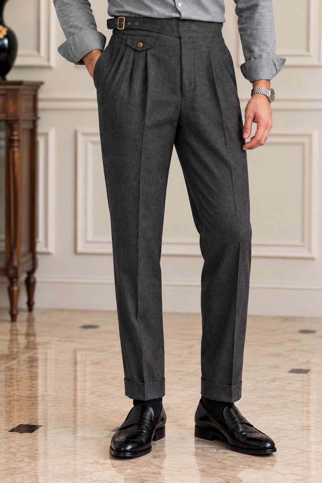 Men's charcoal grey pleated trousers with a high-waisted buckle waistband, tailored fit and cuffed hems, styled with a grey shirt and black leather loafers.