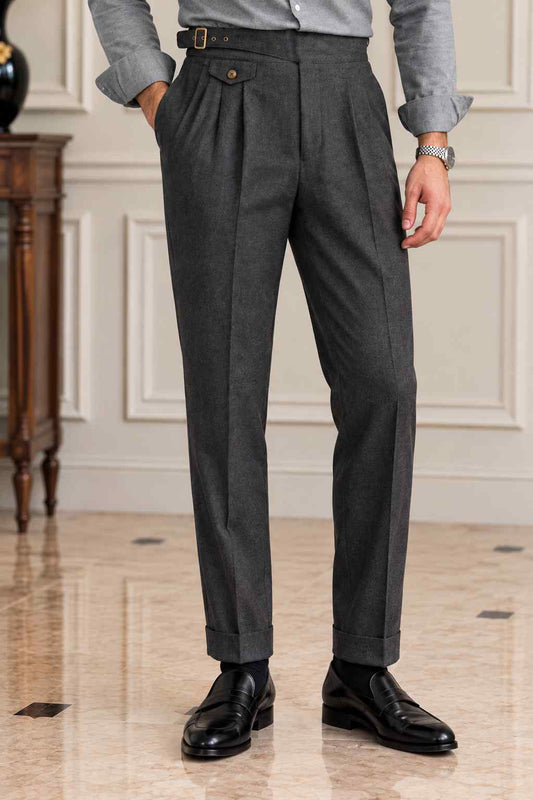 Men's charcoal grey pleated trousers with a high-waisted buckle waistband, tailored fit and cuffed hems, styled with a grey shirt and black leather loafers.