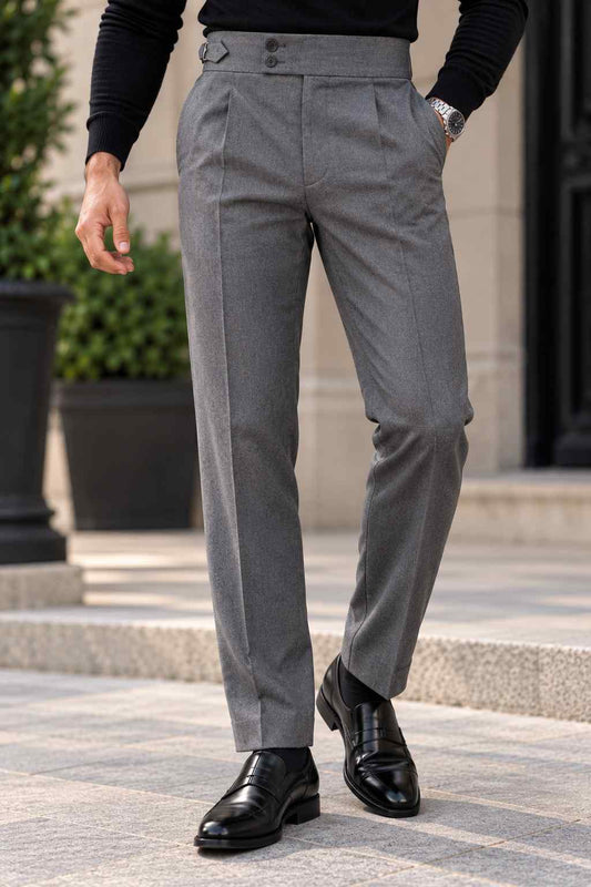 Men's grey high-waisted pleated dress pants with adjustable side buckles and turned-up cuffs, styled with a black turtleneck and black loafers.
