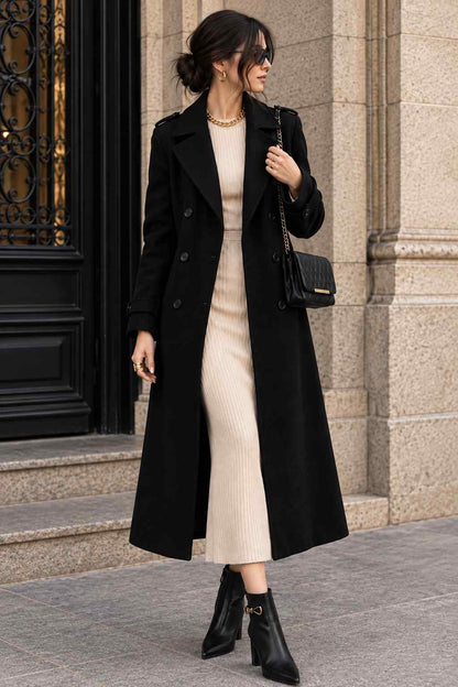 Woman wearing a black double-breasted long coat with wide lapels and shoulder-tab details, styled with a cream dress, black ankle boots, and a chain-strap handbag.
