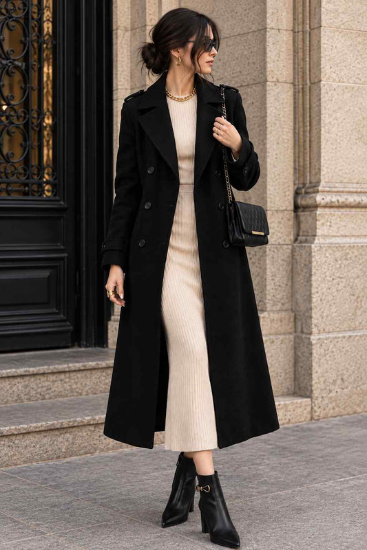 Woman wearing a black double-breasted long coat with wide lapels and shoulder-tab details, styled with a cream dress, black ankle boots, and a chain-strap handbag.